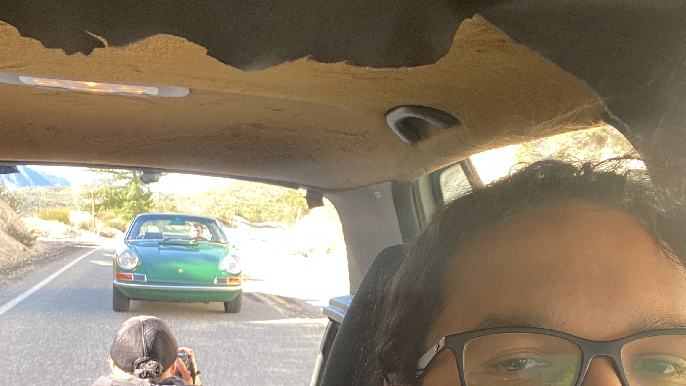 An image facing backwards from the drivers seat of a VW GTI. The headliner is torn, revealing the adhesive underneath. A photographer is taking a photo of a Porsche out of the open rear tailgate.