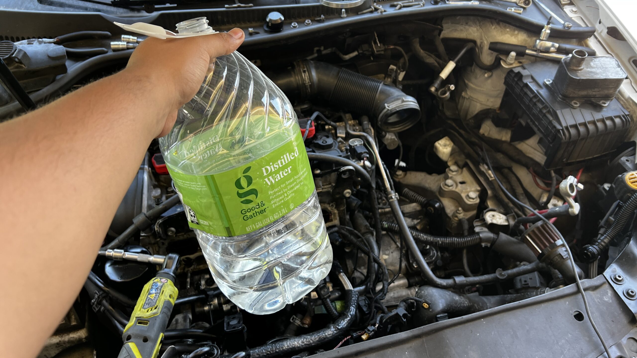 An image of a gallon of distilled water being held above an engine bay. The label on the water gallon is green.