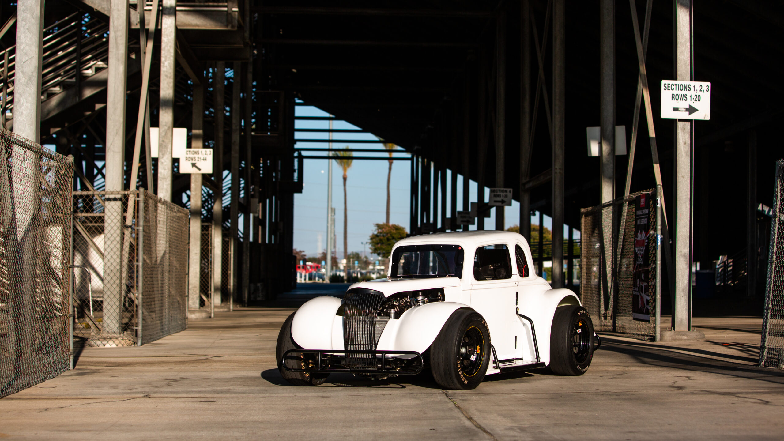 The Legend car parked beneath the grandstands at Irwindale Speedway. The car is white, and the sun is setting behind the camera.