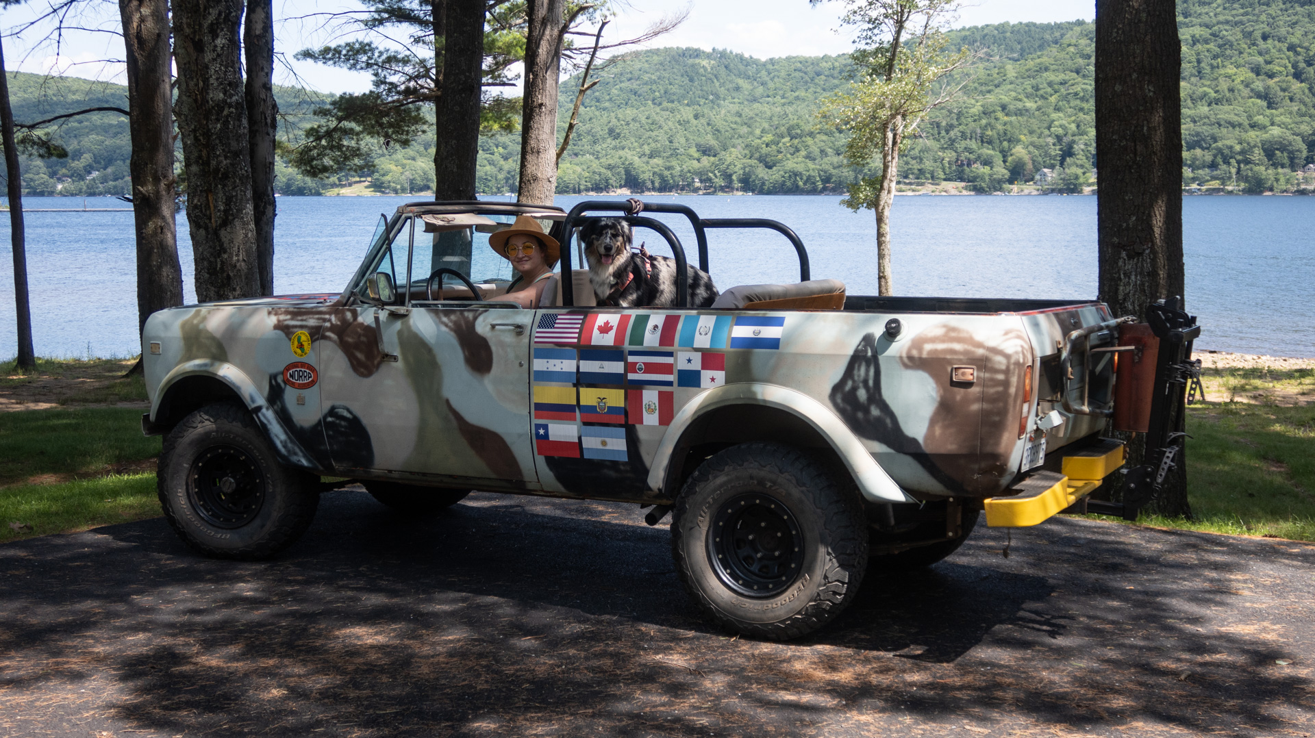 An old truck parked by a lake, woman driving, dog in passenger seat.