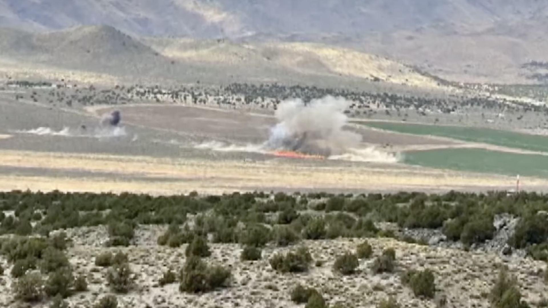 Crash site of fatal Reno Air Races collision. A front of flame can be seen on the desert floor, billowing a large plume of smoke. A second source of smoke can be seen elsewhere.