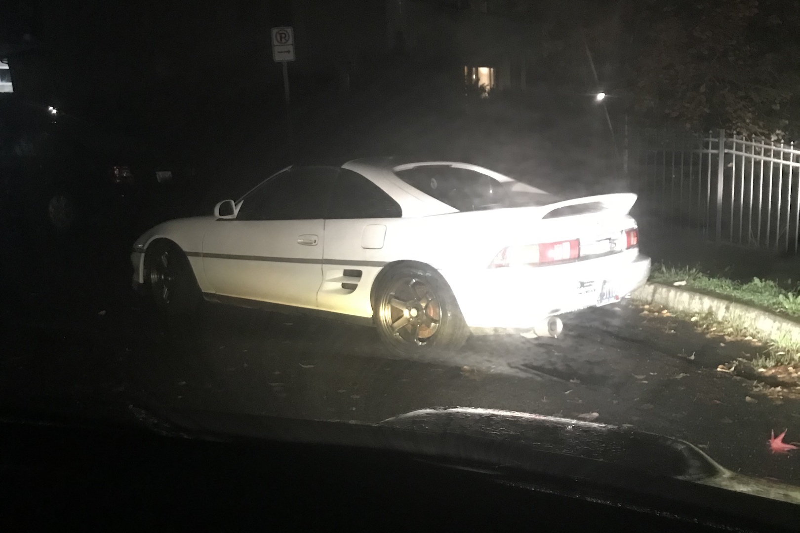 A white sports car, harshly lit by headlights in a dark corner of a neighborhood.