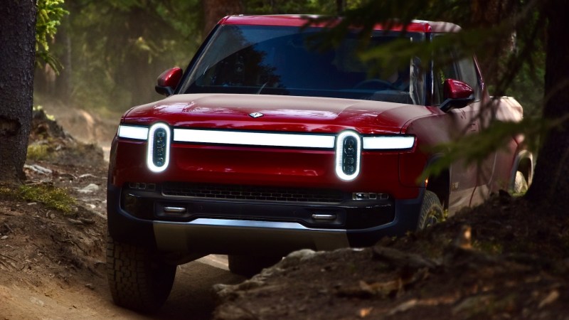 A red Rivian R1T drives down a trail in the woods.