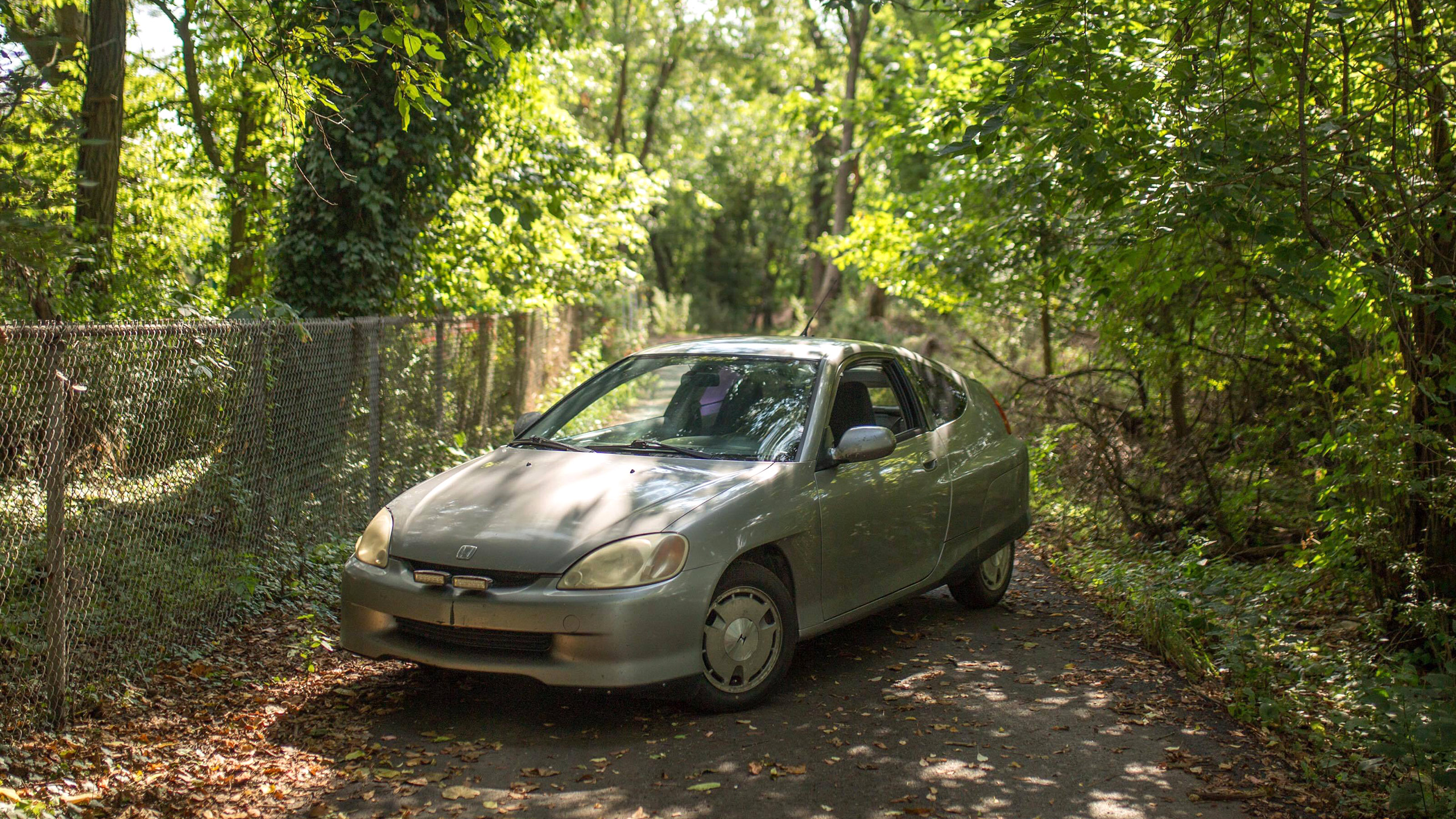 Silver first-gen Honda Insight parked on narrow wooded paved path.