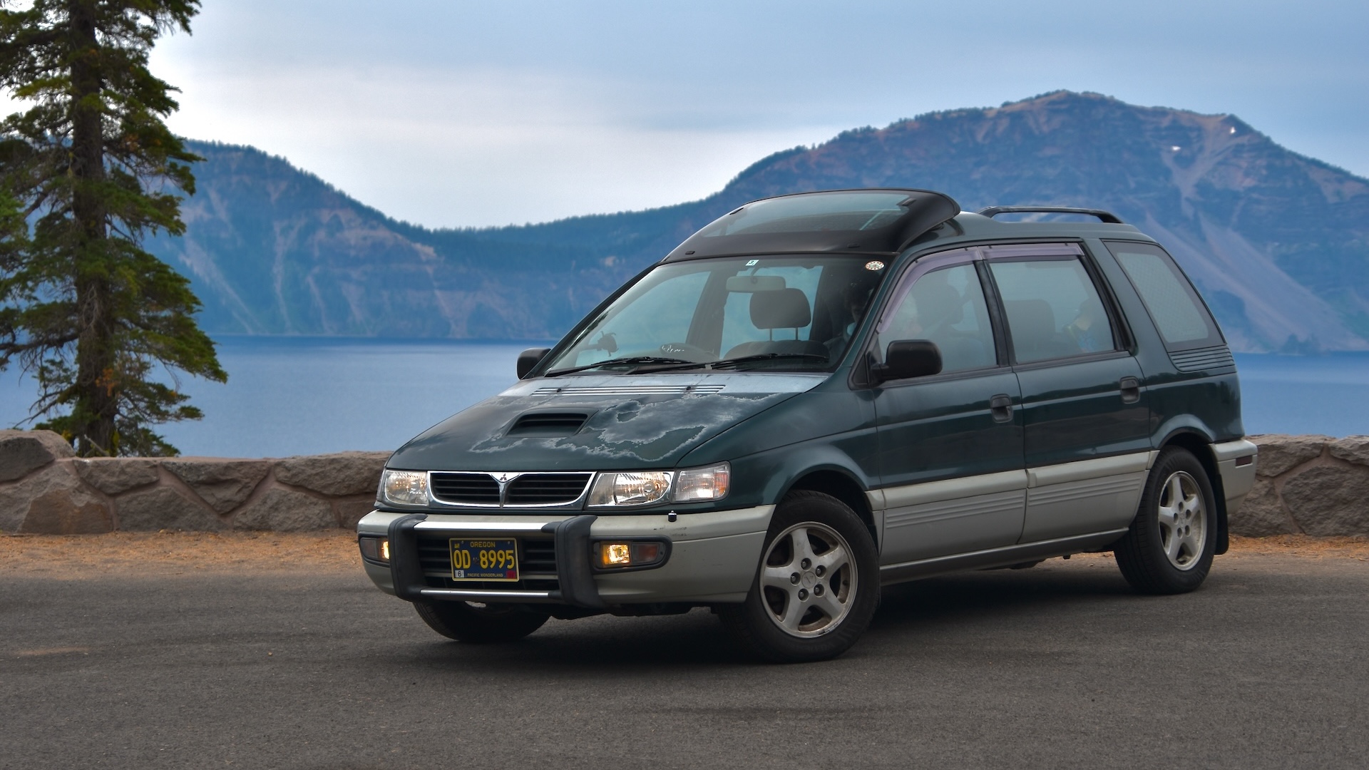 1996 Mitsubishi Chariot Resort Runner GT. A two-tone minivan with a bluish dark green over silver in front of a low cobblestone barrier backed by a conifer. The scene overlooks a grand lake bordered by mountains.