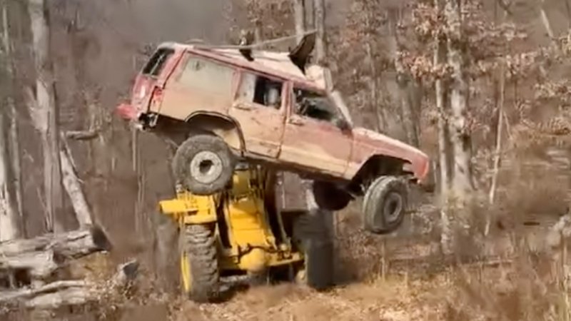 A Jeep Cherokee XJ is carried into the air and moved by a loader, while the loader's forks are going through the SUV's side windows.