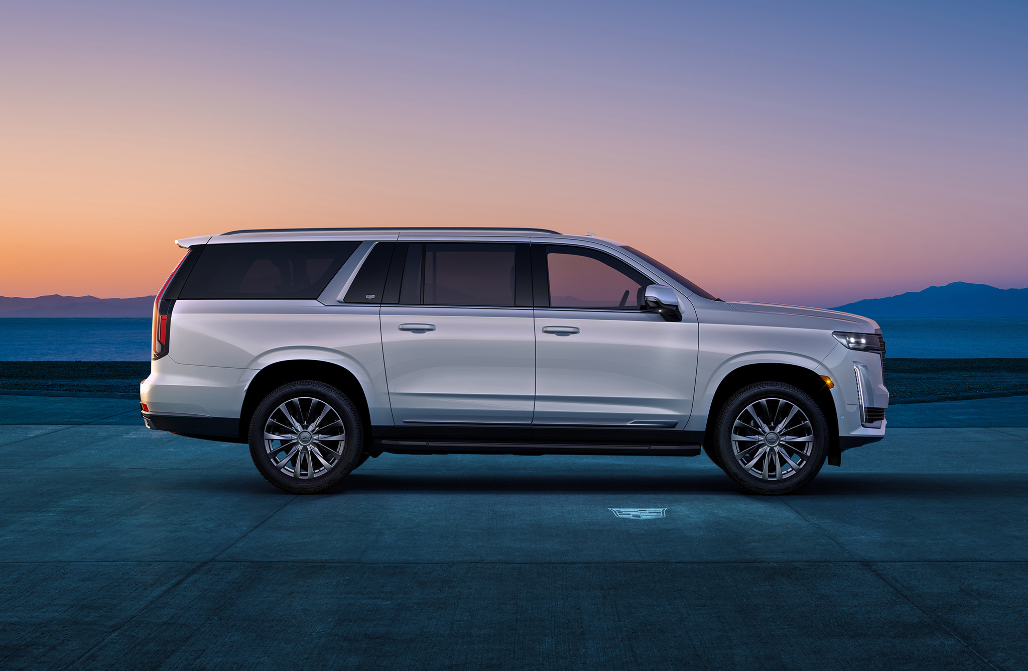 Passenger side profile view of 2023 Escalade ESV Premium Luxury in Crystal White Tricoat with sunset and mountain range in the distance.