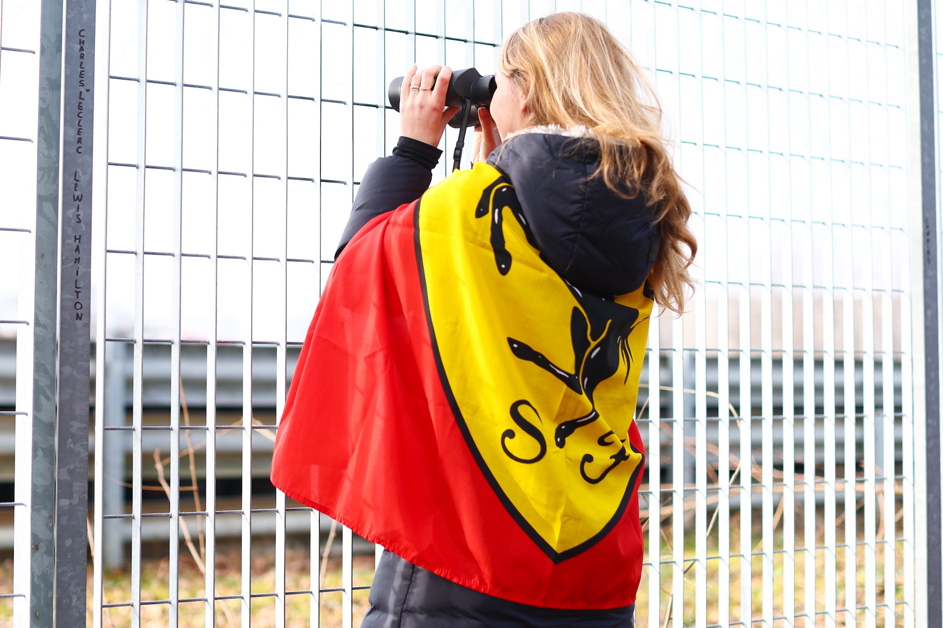 FIORANO MODENESE, ITALY - JANUARY 21: A fan draped in a Ferrari flag holds a pair of binoculars to her eyes, in front of a fence post which has the names of drivers Charles Leclerc and Sir Lewis Hamilton inscribed on it, during Lewis Hamilton's first official days as a Scuderia Ferrari F1 driver at Fiorano Circuit on January 21, 2025 in Fiorano Modenese, Italy. (Photo by Clive Rose/Getty Images)