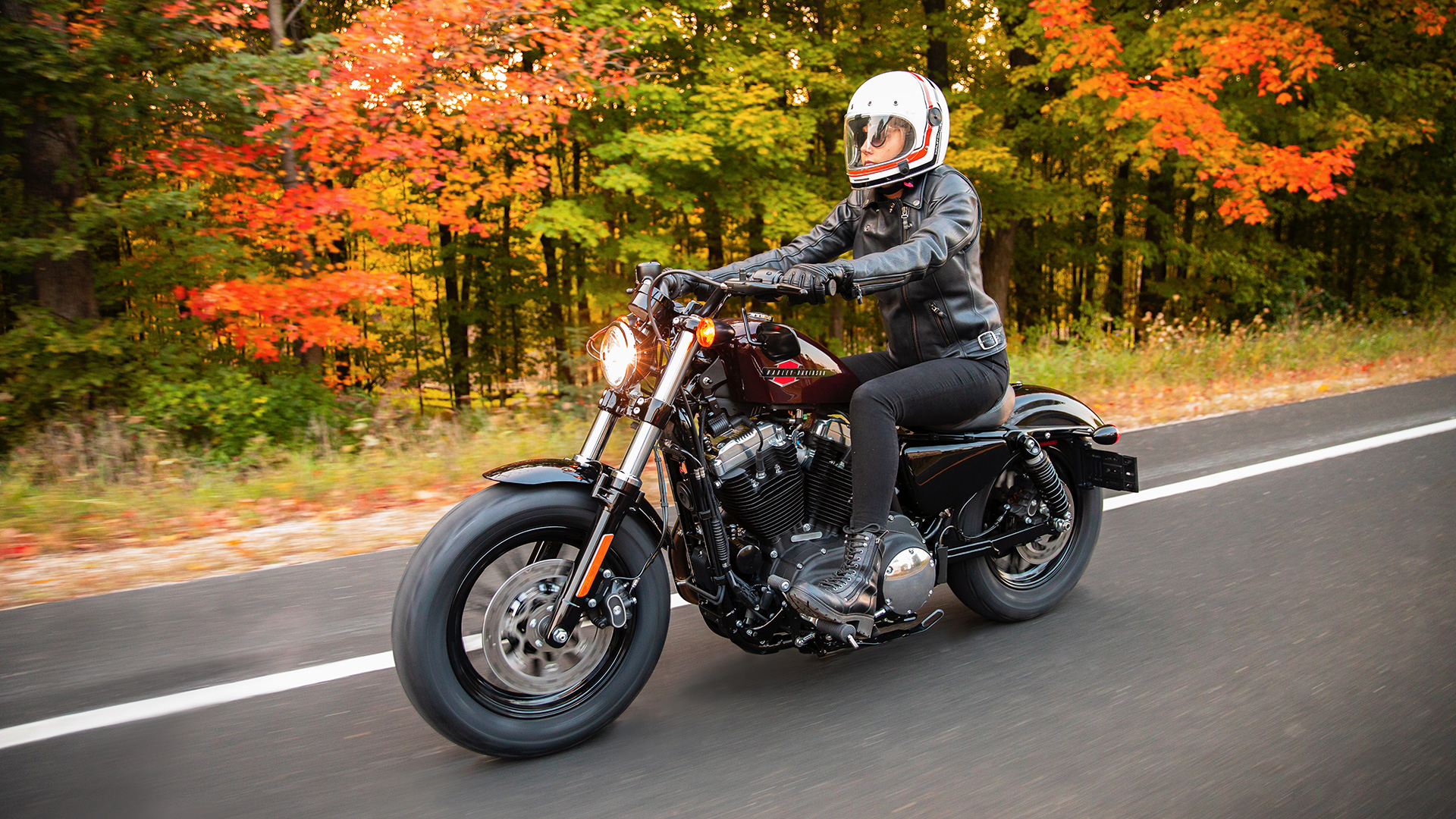 A woman in an upright seating position on a Harley-Davidson as she cruises down a fall road.